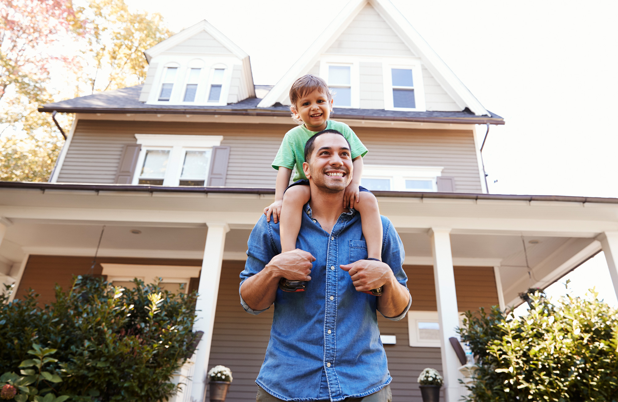 Happy adult with young child on shoulders standing in front of a home